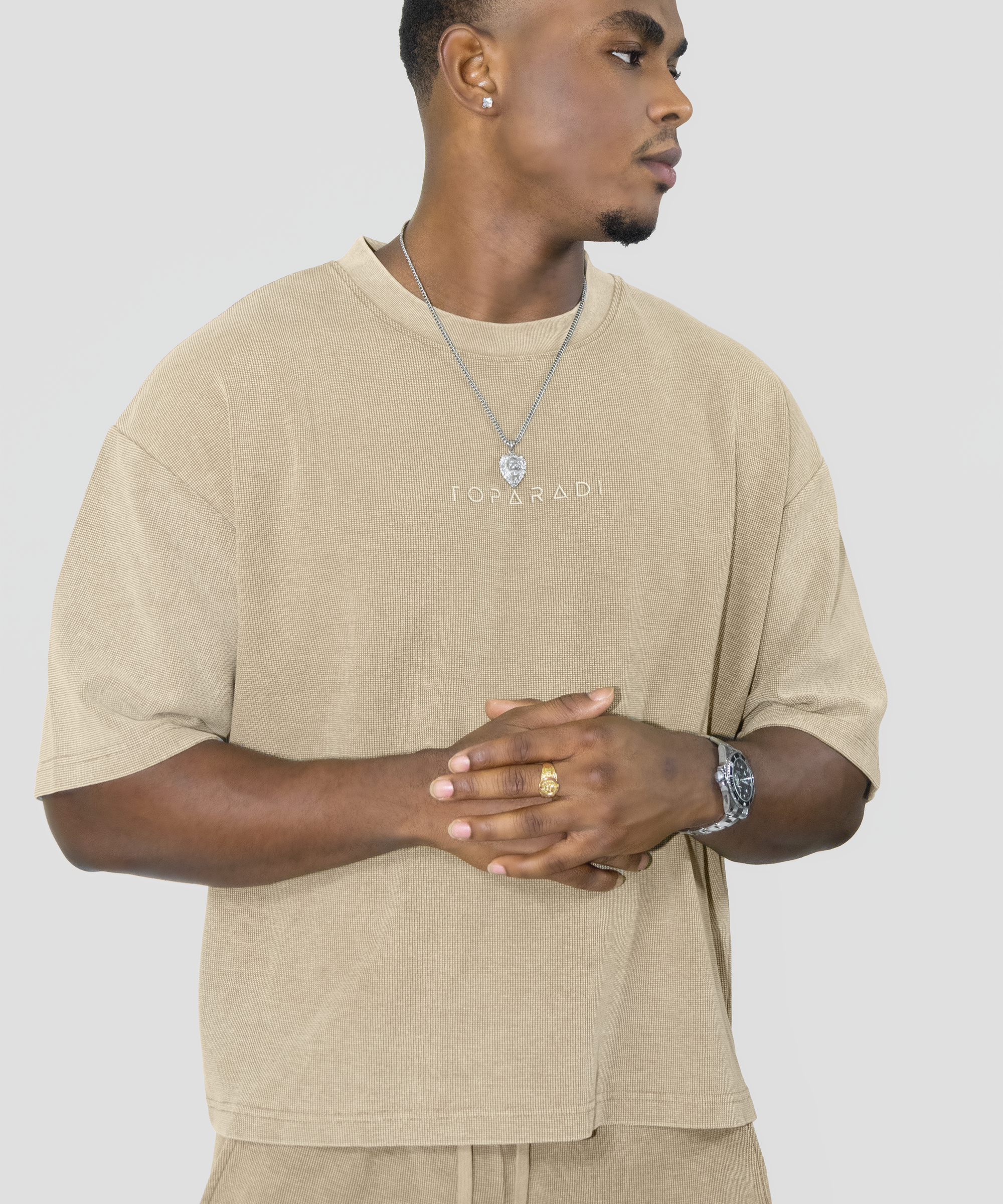 A man wearing a beige oversized t-shirt with 'FOYERADI' printed on the chest, paired with matching beige drawstring pants, against a solid light gray background.