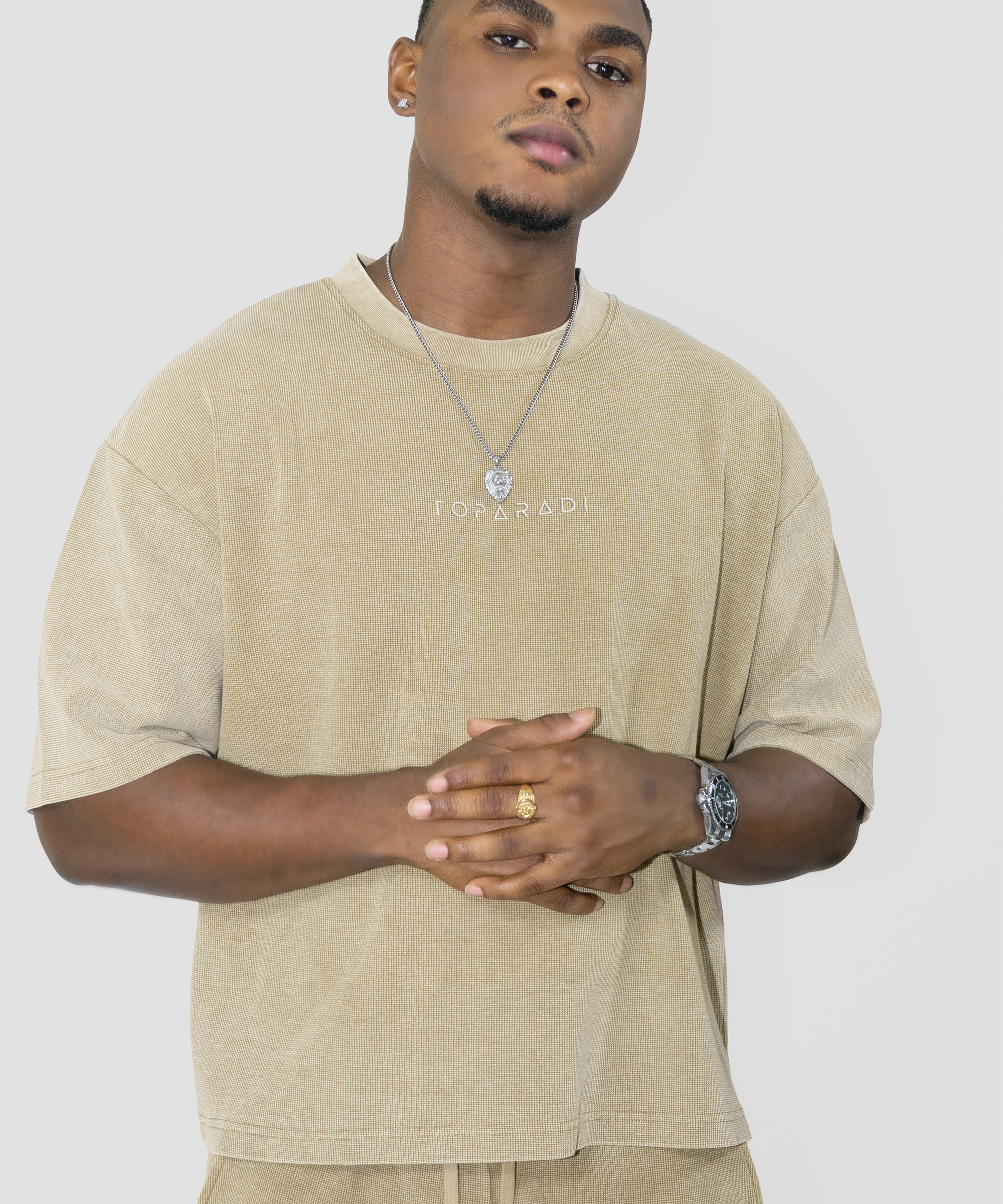 A man wearing a beige oversized t-shirt with 'I'M ON RANI' printed on the chest, paired with matching beige pants, a silver necklace, and a watch, against a light gray background.