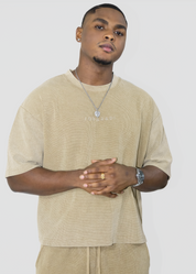 A man wearing a beige oversized t-shirt with 'I'M ON RANI' printed on the chest, paired with matching beige pants, a silver necklace, and a watch, against a light gray background.