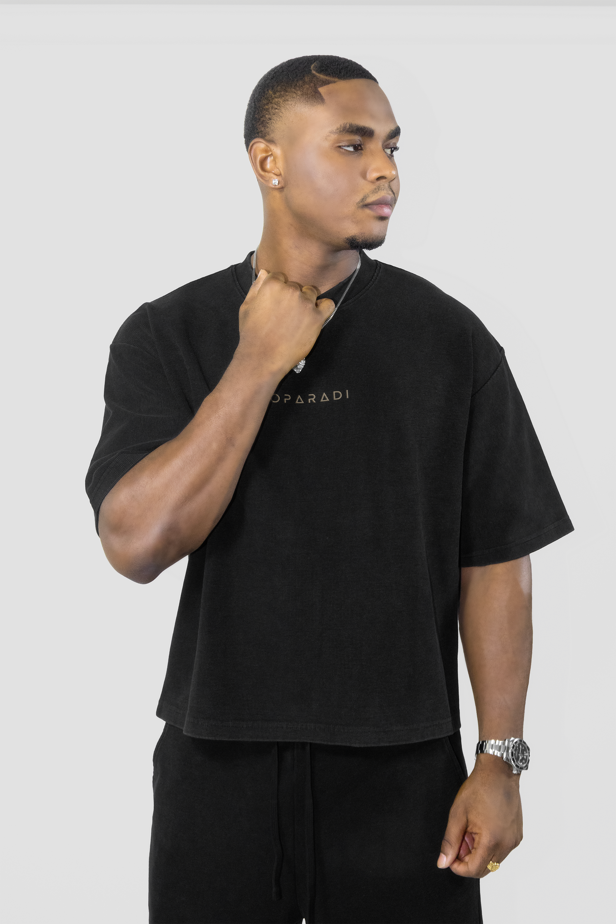 A man wearing a black oversized short-sleeve t-shirt with a small gold logo on the chest, paired with black pants, photographed against a light gray studio background.