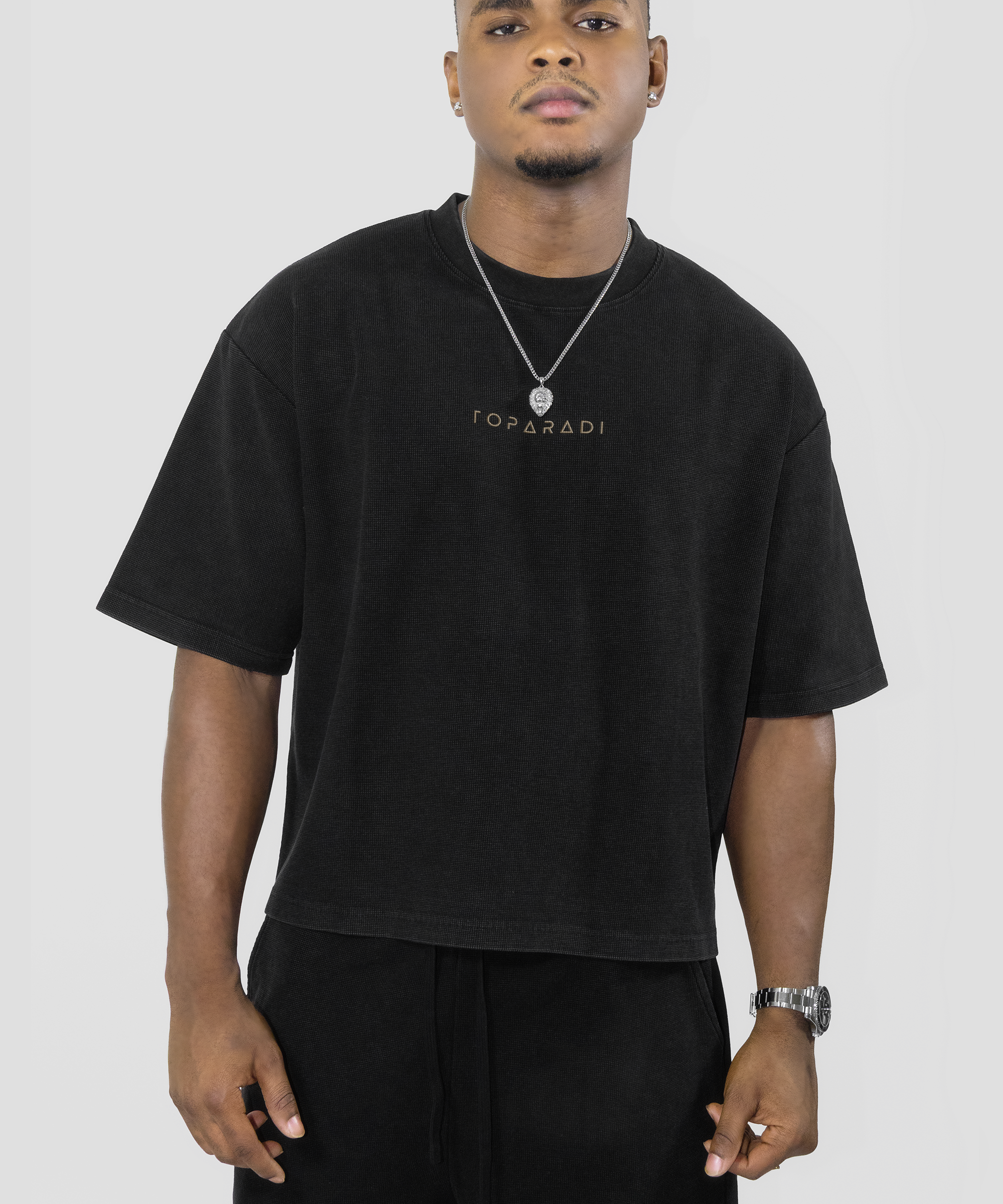 A man wearing a black oversized T-shirt with 'TOPPADI' printed in gold on the chest, paired with black pants, a silver necklace, and a watch, against a light gray studio background.