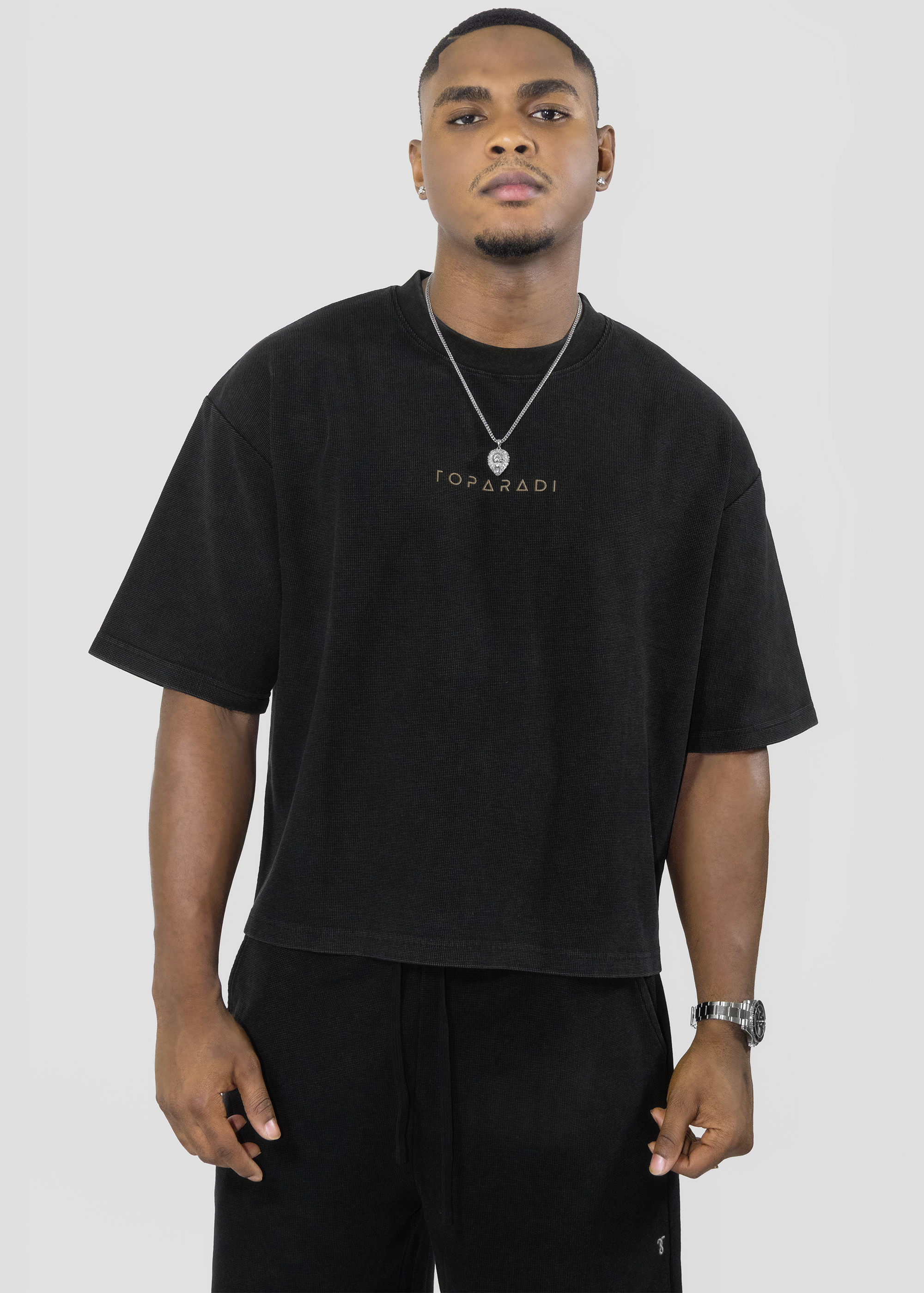 A man wearing a black oversized T-shirt with 'TOPPADI' printed in gold on the chest, paired with black pants, a silver necklace, and a watch, against a light gray studio background.