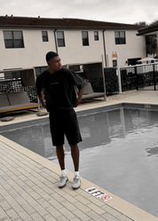A man wearing a black oversized t-shirt and black shorts stands next to a swimming pool at a resort, with a beige building in the background.