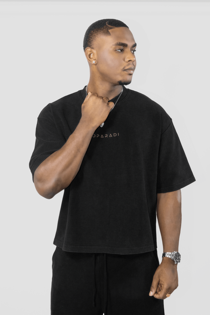 A man wearing a black oversized short-sleeve t-shirt with a small gold logo on the chest, paired with black pants, photographed against a light gray studio background.