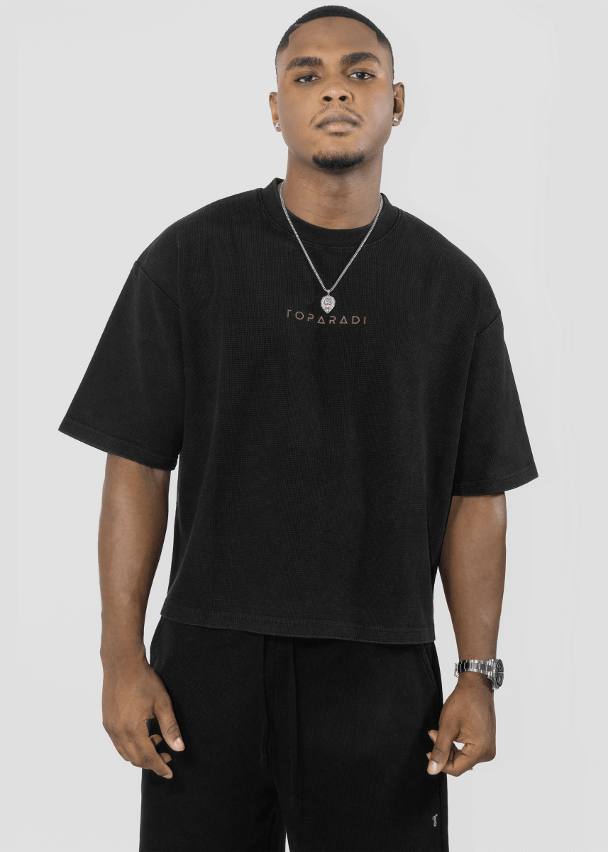 A man wearing a black oversized T-shirt with 'TOPPADI' printed in gold on the chest, paired with black pants, a silver necklace, and a watch, against a light gray studio background.