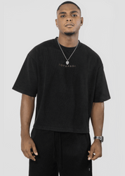 A man wearing a black oversized T-shirt with 'TOPPADI' printed in gold on the chest, paired with black pants, a silver necklace, and a watch, against a light gray studio background.
