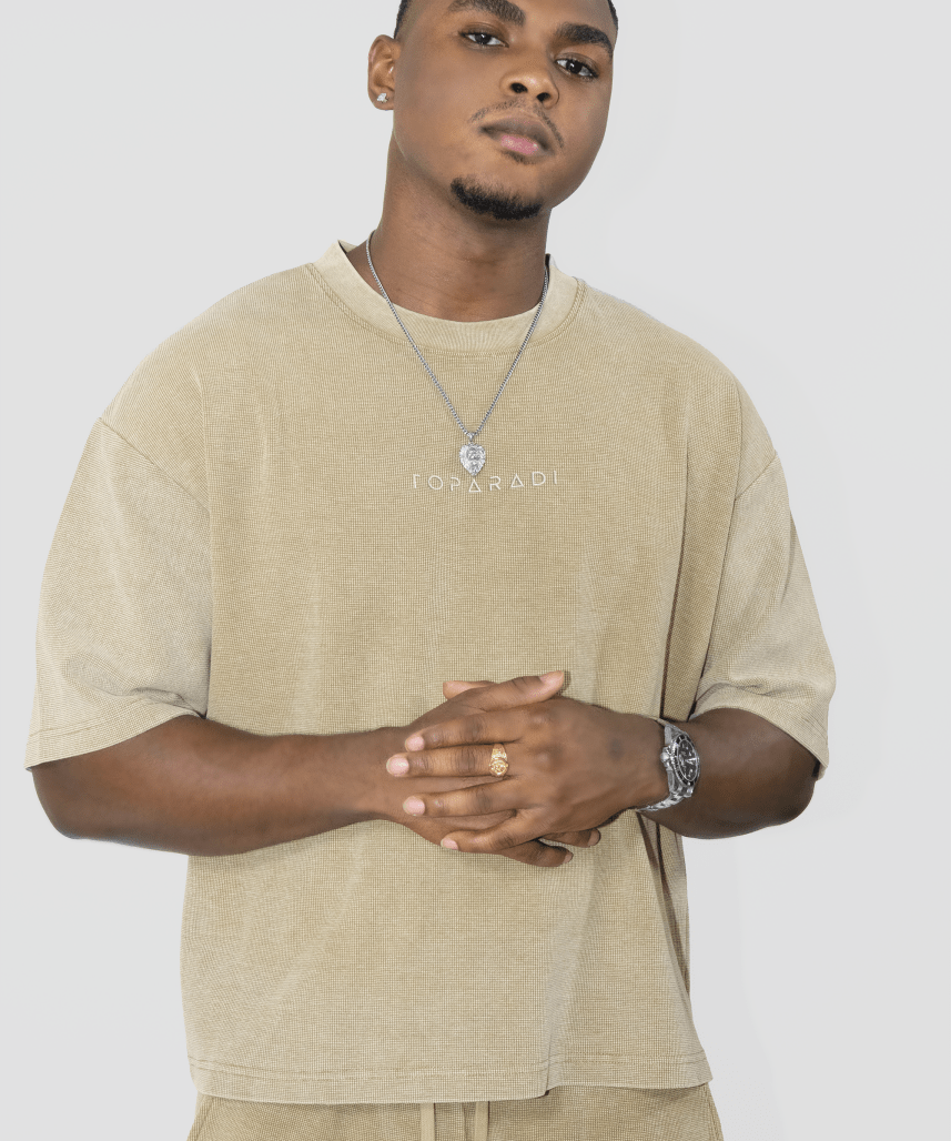 A man wearing a beige oversized t-shirt with 'I'M ON RANI' printed on the chest, paired with matching beige pants, a silver necklace, and a watch, against a light gray background.
