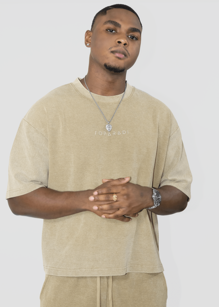 A man wearing a beige oversized t-shirt with 'I'M ON RANI' printed on the chest, paired with matching beige pants, a silver necklace, and a watch, against a light gray background.