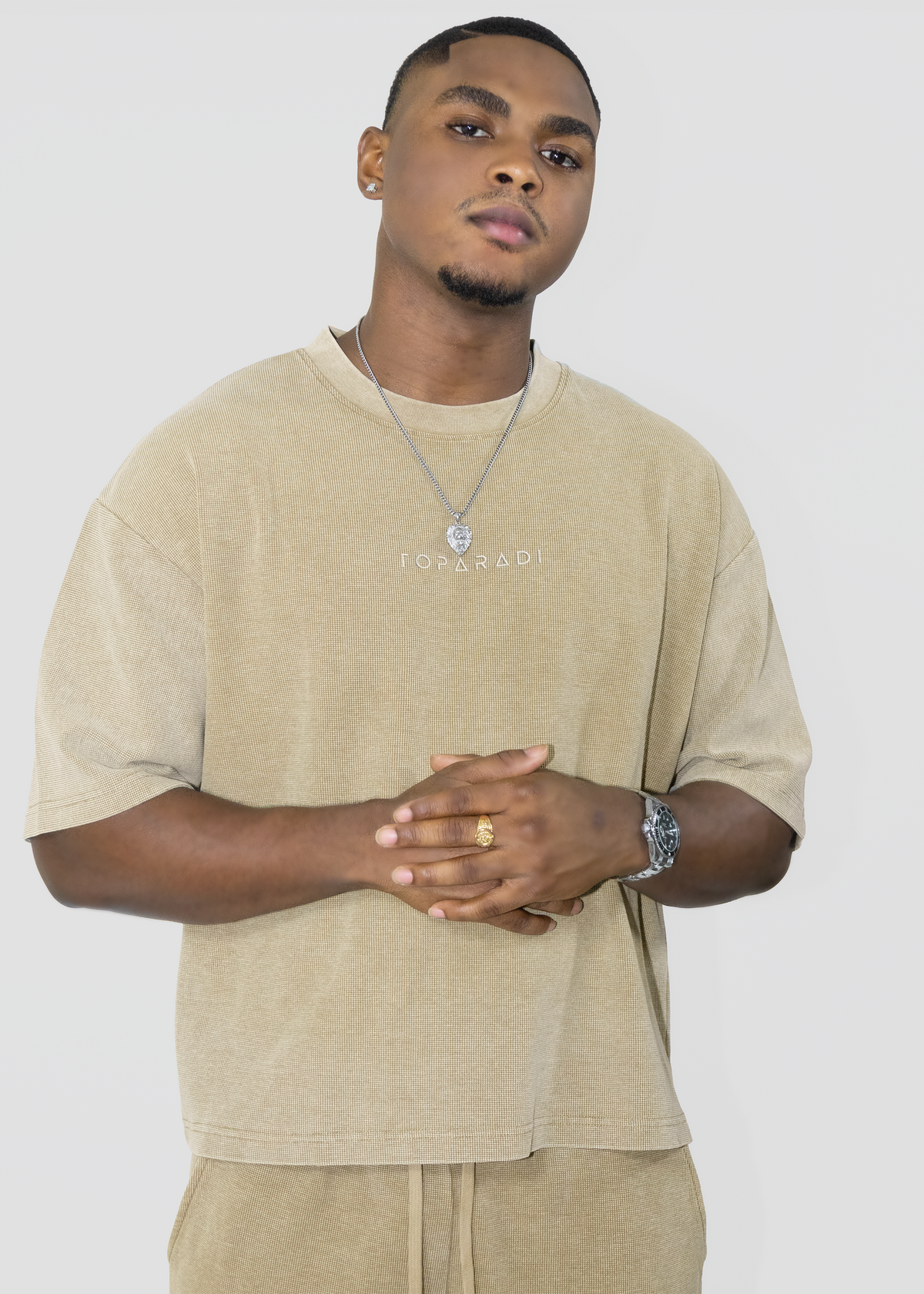 A man wearing a beige oversized t-shirt with 'TOPARADI' printed on the chest, paired with matching beige pants, a silver necklace, and a watch, against a light gray background.