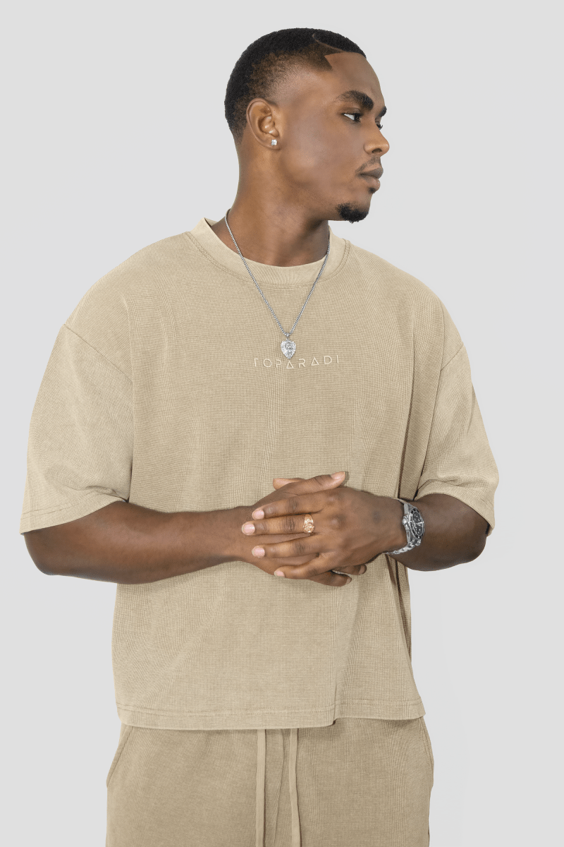 A man wearing a beige oversized t-shirt with 'FOYERADI' printed on the chest, paired with matching beige drawstring pants, against a solid light gray background.