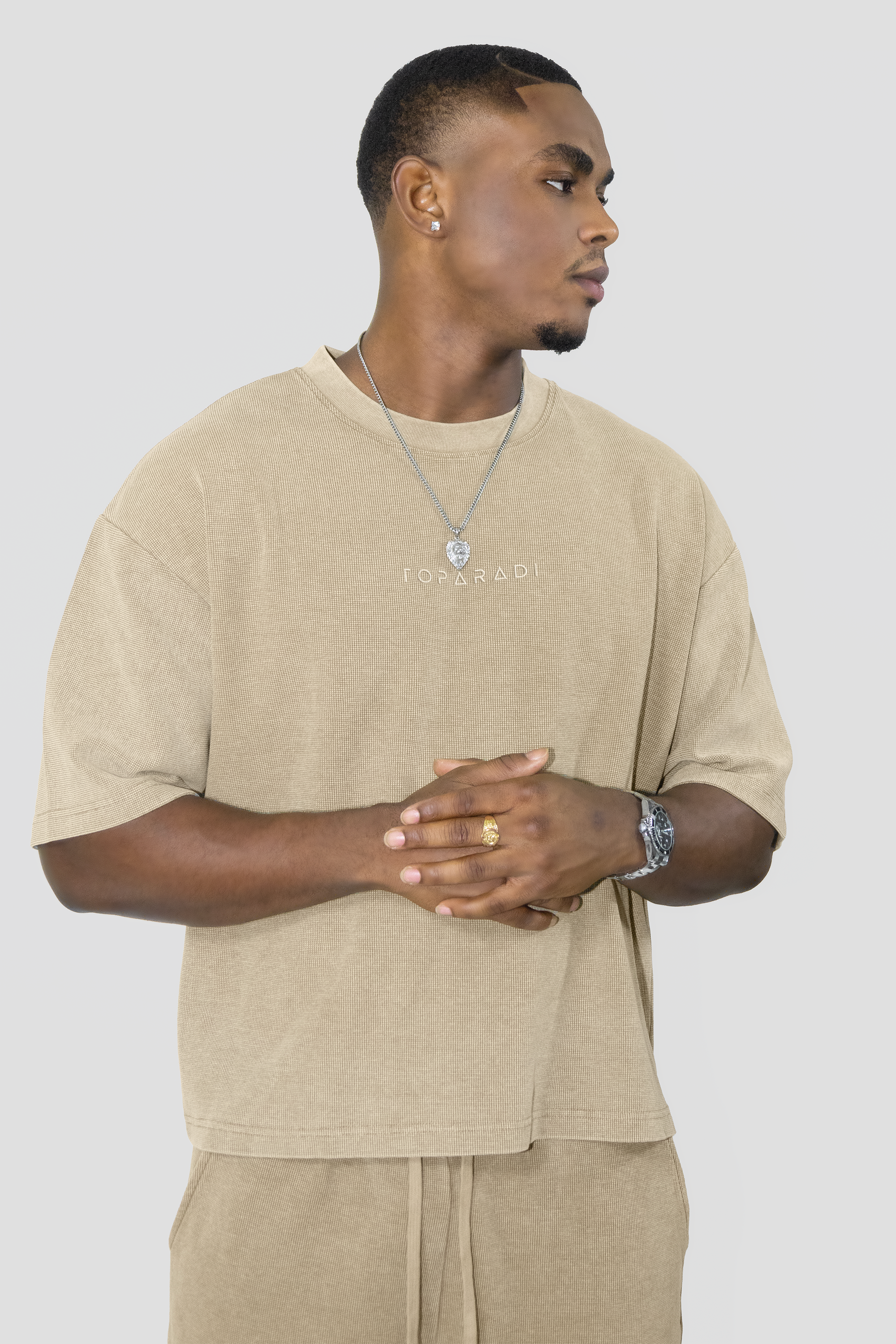 A man wearing a beige oversized t-shirt with 'TOPARADI' printed on the chest, paired with matching beige drawstring pants, against a solid light gray background.