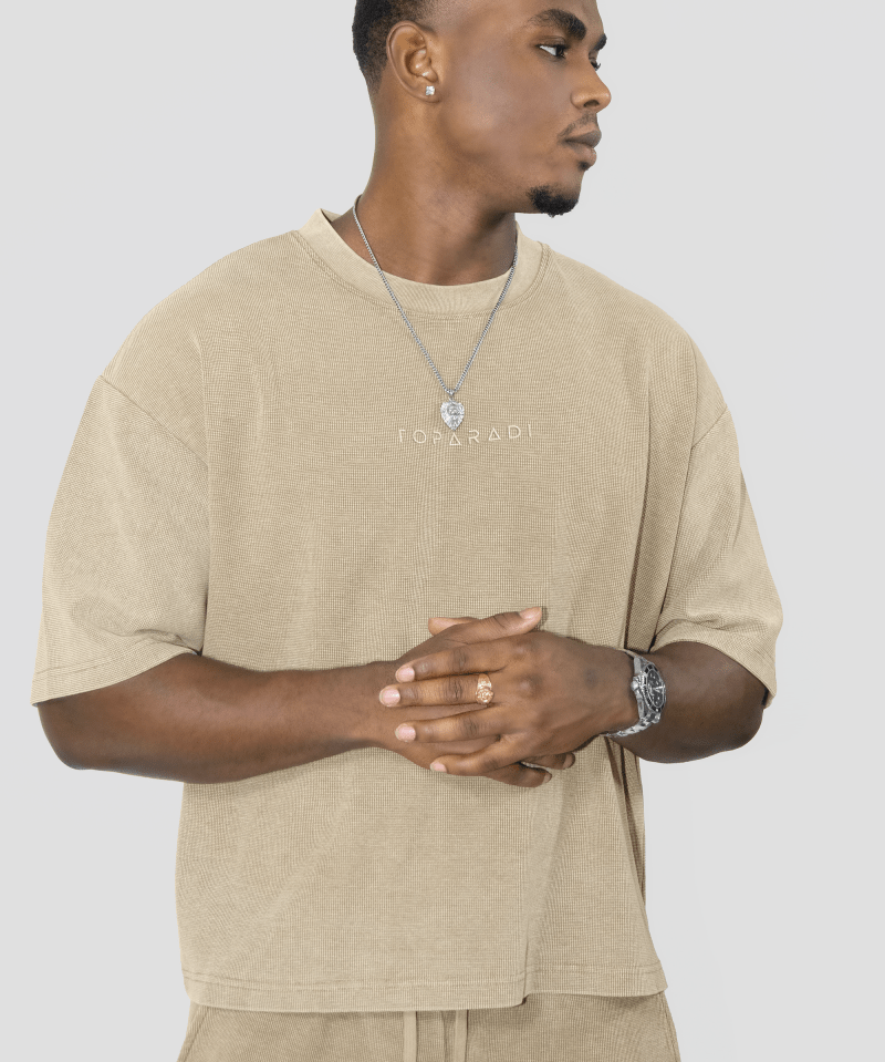 A man wearing a beige oversized t-shirt with 'FOYERADI' printed on the chest, paired with matching beige drawstring pants, against a solid light gray background.