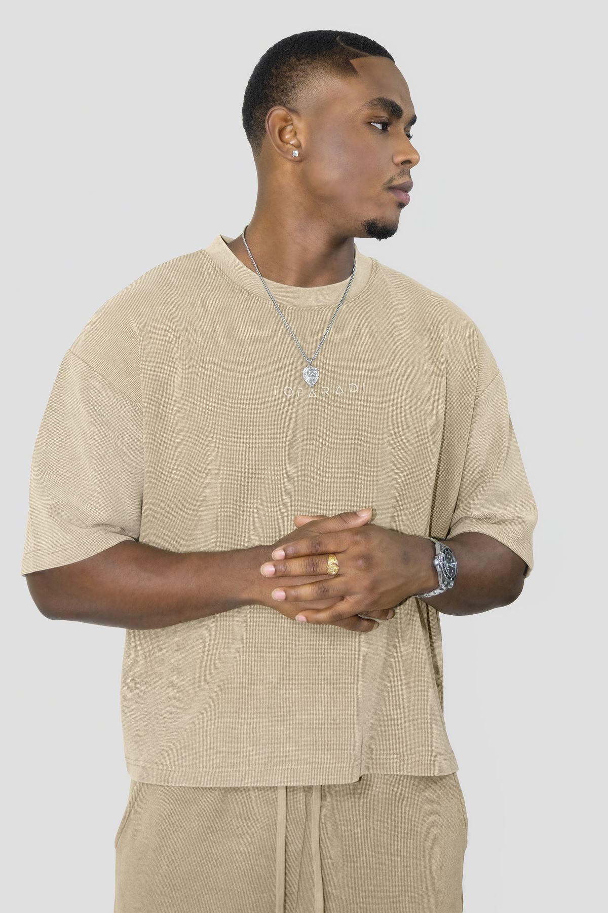 A man wearing a beige oversized t-shirt with 'TOPARADI' printed on the chest, paired with matching beige drawstring pants, against a solid light gray background.