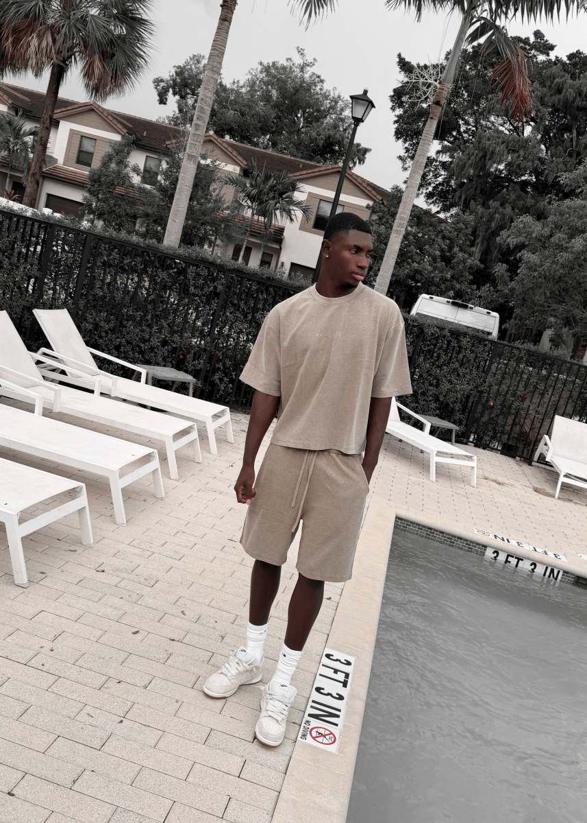 A man stands by a swimming pool wearing a beige short-sleeve crewneck t-shirt and matching beige shorts, with white socks and white sneakers. White lounge chairs, palm trees, and resort-style buildings are visible in the background.