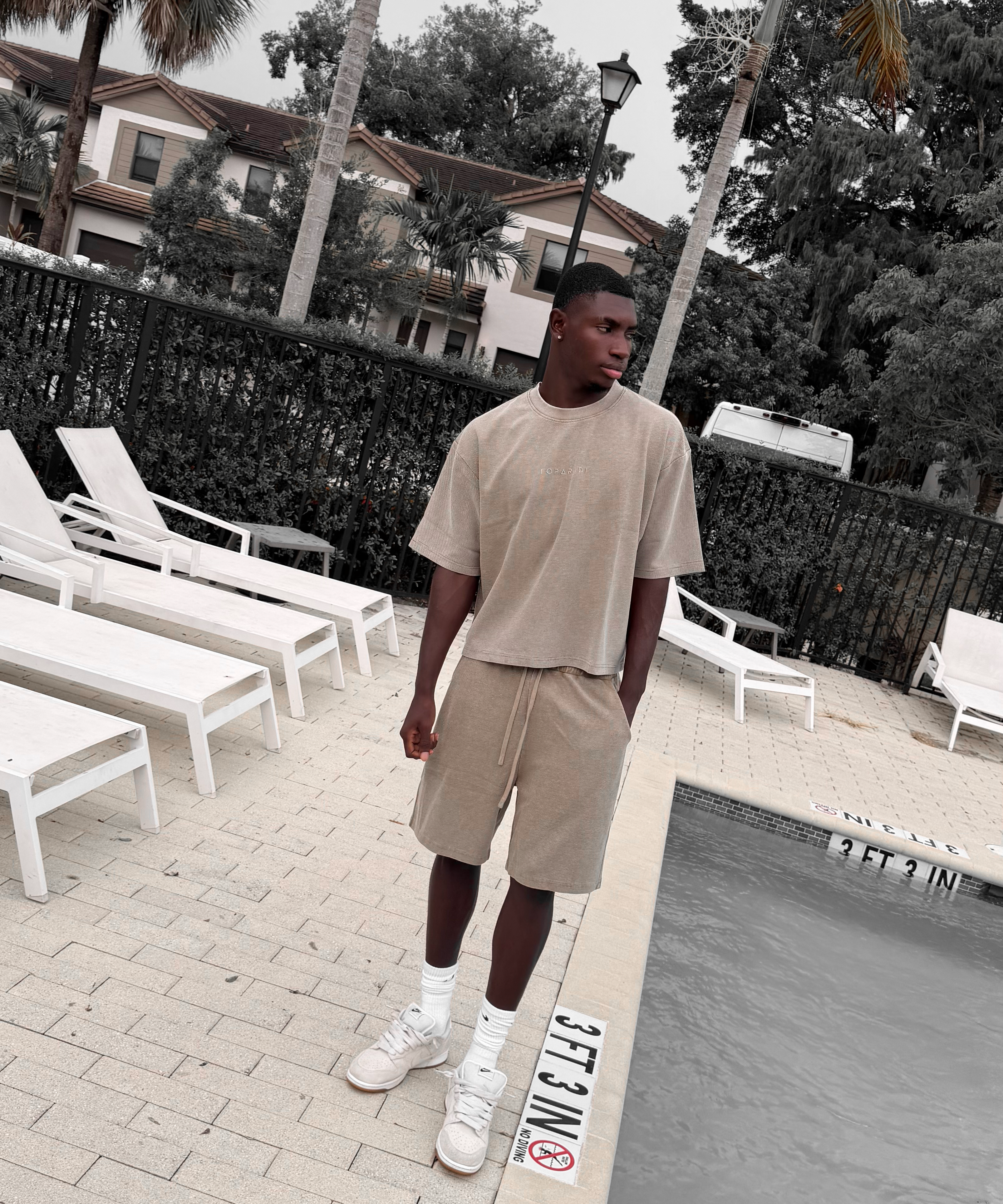 A man stands by a swimming pool wearing a beige short-sleeve crewneck t-shirt and matching beige shorts, with white socks and white sneakers. White lounge chairs, palm trees, and resort-style buildings are visible in the background.