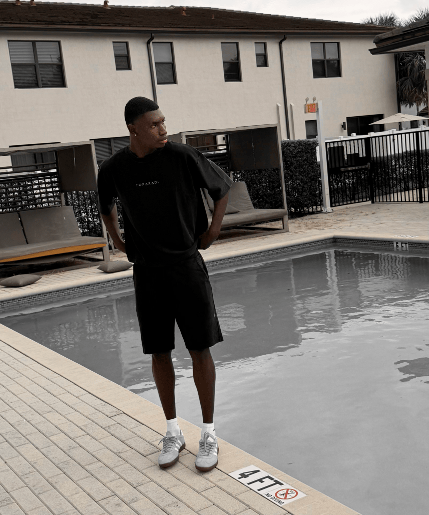 A man wearing a black oversized t-shirt and black shorts stands next to a swimming pool at a resort, with a beige building in the background.