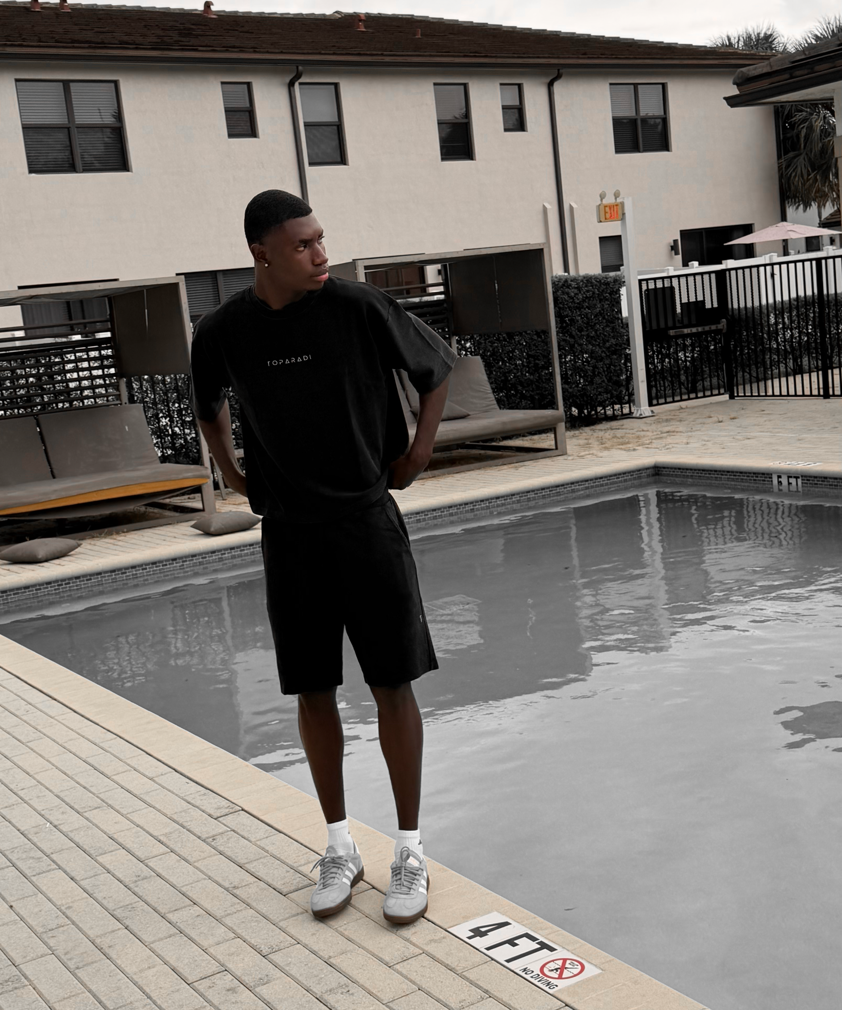 A man wearing a black oversized t-shirt and black shorts stands next to a swimming pool at a resort, with a beige building in the background.